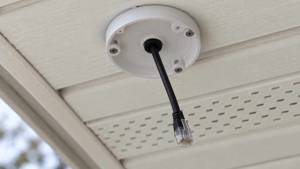A white round camera base screwed to the underside of the eave, a black network cable hanging straight down from a rubber grommet in its center and ending in a clear plug, a vented soffit panel behind it